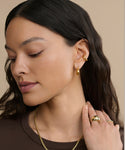 A woman with long dark hair wears gold hoop earrings, a gold necklace, and the Felix Dome Ring by Jenni Kayne Jewelry. She is looking down, touching her neck against a beige background.