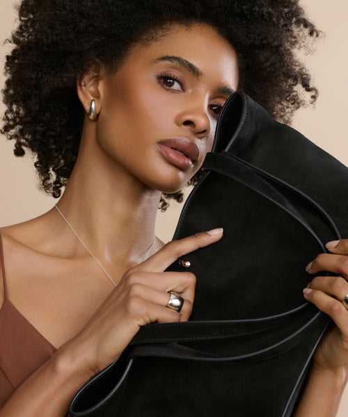 A woman with curly hair holds a large black suede clutch near her face. She wears a brown top, silver hoops, a delicate necklace, and the Felix Dome Ring, posing against a neutral background.