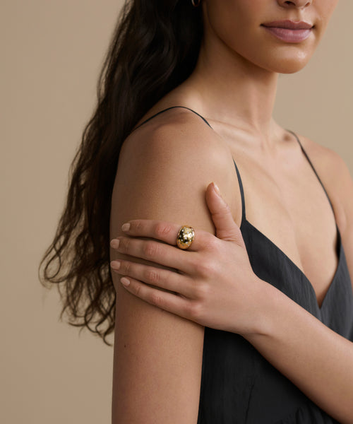 A woman in a black sleeveless dress rests her hand on her upper arm, showing off the large, round Diamond Felix Dome Ring. Her long wavy hair falls over one shoulder, set against a beige background.