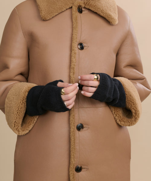 A person shown from the shoulders down wears a tan shearling coat with large buttons, several gold rings, and black Fingerless Cashmere Gloves.