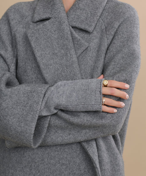 A person in a cozy gray wool coat and Fingerless Cashmere Gloves crosses their arms, displaying manicured nails and gold rings against a neutral beige background.
