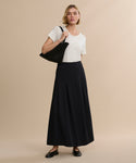 A woman with short blonde hair and a neutral expression stands against a beige background in a white t-shirt, black flats, and a black shoulder bag, wearing the Florence Skirt—a long black crepe midi skirt.