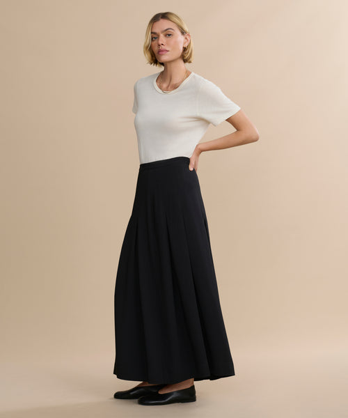 A person poses against a beige background wearing a white short-sleeve top, the Florence Skirt in black as a long midi skirt, and black flats, with one hand on their hip.