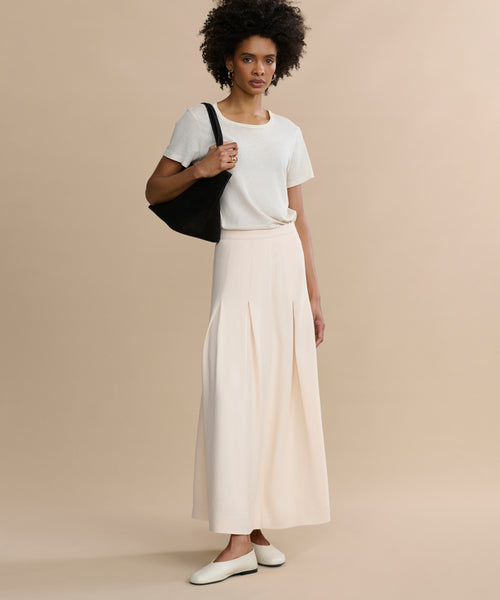 A woman stands against a beige background wearing a white short-sleeve t-shirt, the Florence Skirt in cream crepe, white flats, and carrying a black shoulder bag.