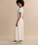 A woman with short curly hair stands sideways against a beige background, wearing a white short-sleeve t-shirt, white slip-on shoes, and the Florence Skirt—a long cream crepe skirt.