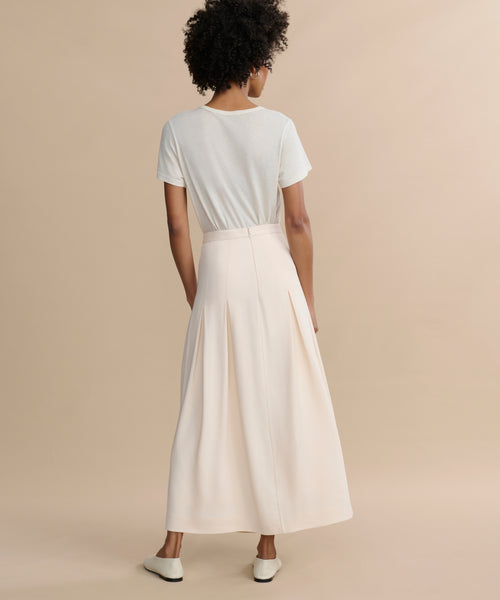 Facing away, a person with short curly hair wears the Florence Skirt—a high-waisted, cream crepe midi skirt with pleats—paired with a short-sleeve white t-shirt and white shoes against a plain beige background.