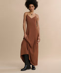 A person with short curly hair wears the Florence Slip Dress in brown crepe with thin straps and black boots, standing against a beige background and gently holding one side of the dress.