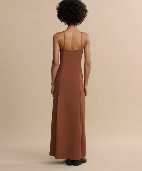A person with curly hair stands against a beige background, seen from behind in a long brown Florence Slip Dress featuring thin straps and a fitted bodice, crafted from crepe fabric, paired with black shoes.
