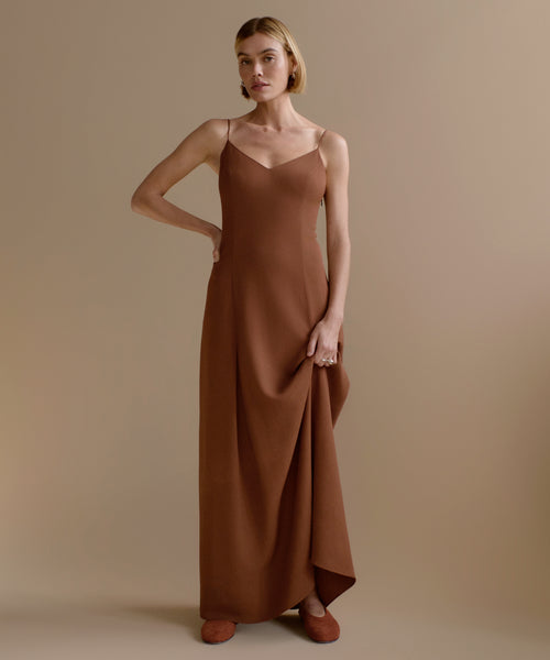 A woman with short blonde hair models the Florence Slip Dress, featuring thin straps and a fitted bodice in brown crepe, posing with one hand on her hip against a plain beige background.