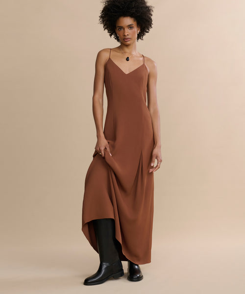 A person with short curly hair wears the Florence Slip Dress in brown crepe with thin straps and black boots, standing against a beige background and gently holding one side of the dress.