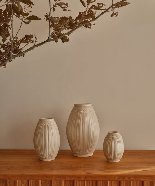 A handcrafted Porta Ceramic Vase and two other beige ceramic vases with vertical ridges sit on a wooden surface beneath a branch with dark leaves, set against a plain beige wall.