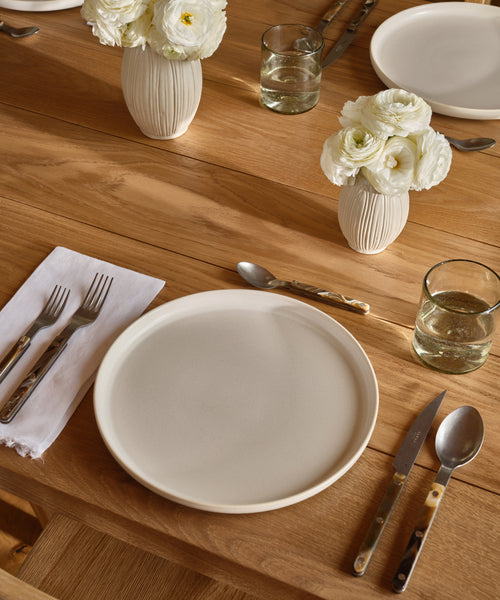 A wooden table set with a white ceramic plate, cutlery, a white napkin, two glasses of water, and two Porta Ceramic Vases filled with white flowers.