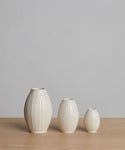 Three cream-colored Porta Ceramic Vases of different sizes, featuring vertical ridges, are arranged in a row on a light wooden surface with a plain gray background.