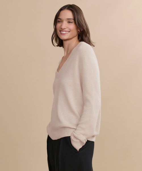 A woman with shoulder-length brown hair smiles against a beige background, wearing the Flynn Cashmere Sweater in light beige with a V-neck and black pants, her hands casually in her pockets.