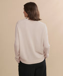 A person with shoulder-length brown hair, seen from behind against a beige background, is wearing the Flynn Cashmere Sweater in light pink paired with black pants.