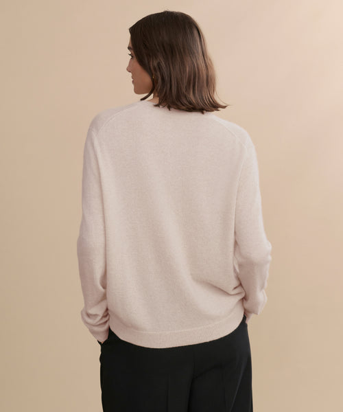 A person with shoulder-length brown hair, seen from behind against a beige background, is wearing the Flynn Cashmere Sweater in light pink paired with black pants.