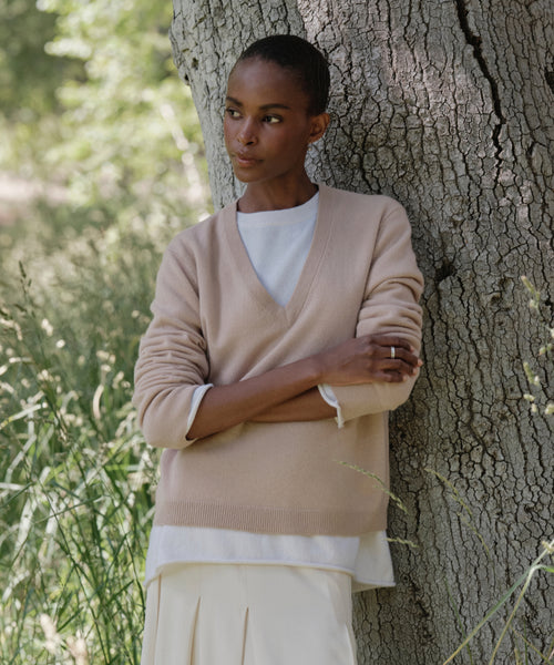 A person stands outdoors, leaning against a tree in the Flynn Cashmere Sweater, a beige V-neck style layered over a white shirt and cream skirt, with sunlight streaming through lush greenery in the background.