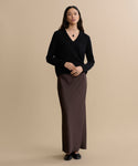 A woman with long dark hair stands against a beige background, wearing the Flynn Cashmere Sweater in a V-neck style, paired with a long brown skirt and black shoes. She accessorizes with a pendant necklace and faces the camera with hands lightly clasped.
