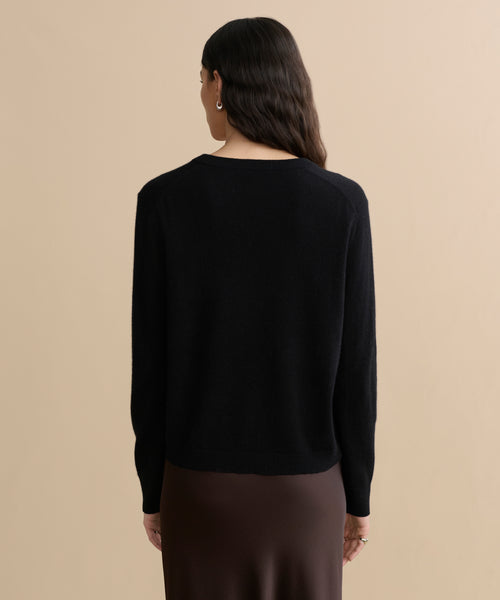 A person with long wavy hair stands facing away, wearing the Flynn Cashmere Sweater in black and a dark brown skirt against a plain beige background.