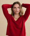 A woman with light brown hair and a neutral expression poses with her arms raised behind her head, wearing the Flynn Cashmere Sweater in relaxed red v-neck knit and a gold necklace against a beige background.