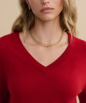A woman wears the Flynn Cashmere Sweater, a relaxed-fit red V-neck knit, paired with layered gold necklaces and hoop earrings. She has wavy blonde hair, a neutral expression, and stands against a beige background.