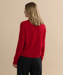 A person with shoulder-length blonde hair, wearing the Flynn Cashmere Sweater in bright red and black pants, stands facing away from the camera against a plain beige background.