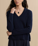 A woman with long wavy hair wears the Flynn Cashmere Sweater in navy blue, paired with dark pants, a black teardrop pendant necklace, and gold hoop earrings. She lightly touches her chin with one hand while crossing her other arm.