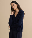 A woman with long, wavy dark hair wears the Flynn Cashmere Sweater in navy and dark pants, accessorized with a necklace and rings. She stands against a beige background, looking relaxed and thoughtful.