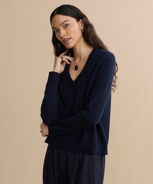 A woman with long, wavy dark hair wears the Flynn Cashmere Sweater in navy and dark pants, accessorized with a necklace and rings. She stands against a beige background, looking relaxed and thoughtful.