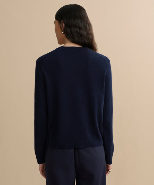 A person with wavy dark hair stands with their back to the camera, wearing a navy blue Flynn Cashmere Sweater and dark pants against a plain beige background.
