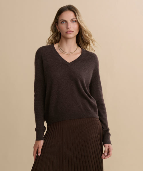 A woman with wavy blonde hair wears the Flynn Cashmere Sweater in dark brown, styled with a matching pleated skirt. She stands against a beige background, accessorized minimally and maintaining a neutral expression.