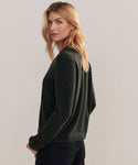 A woman with blonde hair stands sideways, looking over her shoulder at the camera, wearing the Flynn Cashmere Sweater in dark green and black pants against a plain light background.