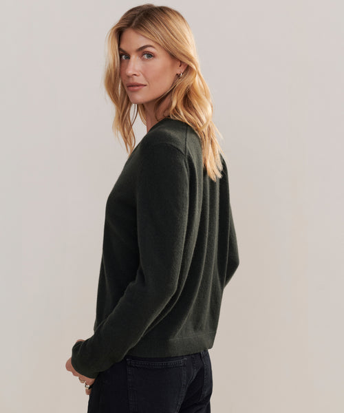 A woman with blonde hair stands sideways, looking over her shoulder at the camera, wearing the Flynn Cashmere Sweater in dark green and black pants against a plain light background.