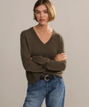 A woman with short, light brown hair wears the Flynn Cashmere Sweater in a relaxed fit with a V-neck, paired with blue jeans, a black belt, and gold jewelry as she stands against a plain light background with arms crossed.