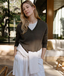 Wearing the Flynn Cashmere Sweater, a woman with long blonde hair poses indoors with hands in her pockets; she pairs the dark v-neck sweater over a white shirt, tying another white sweater around her waist in a relaxed, modern setting.