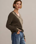 Woman with short brown hair wears the Flynn Cashmere Sweater, a relaxed fit knit V-neck in khaki, paired with blue jeans, standing with one hand in her pocket against a plain light background.