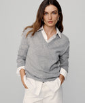 A woman with long brown hair wears the Flynn Cashmere Sweater—a relaxed fit gray knit—over a white collared shirt and white pants, standing against a plain light gray background with her hands in her pockets.