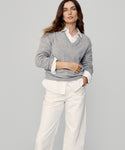 A woman stands against a plain background, wearing the Flynn Cashmere Sweater in light gray over a white collared shirt and wide-leg white pants. She has long brown hair, a neutral expression, and one hand in her pocket.