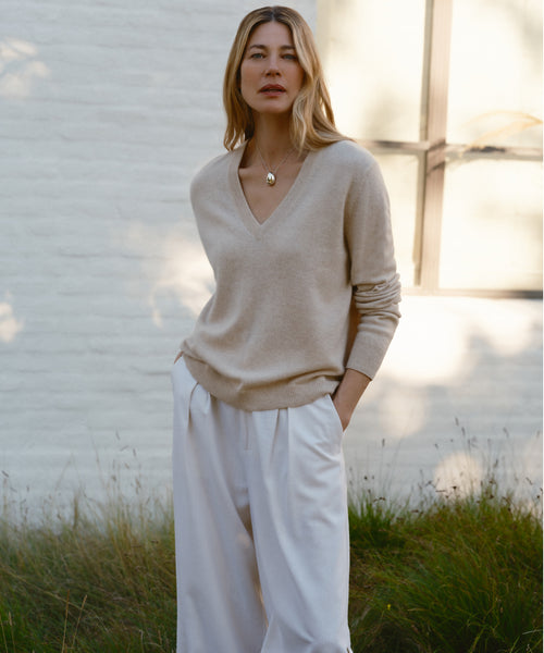 A woman with light brown hair stands outside in front of a white brick wall and window, wearing the beige Flynn Cashmere Sweater, white wide-leg pants, and a pendant necklace, with her hands in her pockets.