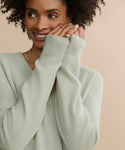 A woman with curly hair smiles gently, wearing the Flynn Cashmere Sweater in light sage green with a relaxed fit and long sleeves, paired with a gold ring on her finger, posed against a beige background.