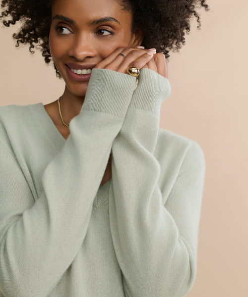 A woman with curly hair smiles gently, wearing the Flynn Cashmere Sweater in light sage green with a relaxed fit and long sleeves, paired with a gold ring on her finger, posed against a beige background.