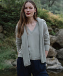 A woman with long brown hair stands outdoors by rocks and greenery, wearing the Flynn Cashmere Sweater in light gray, paired with a matching sweater draped over her shoulders and dark blue pants. She appears calm.