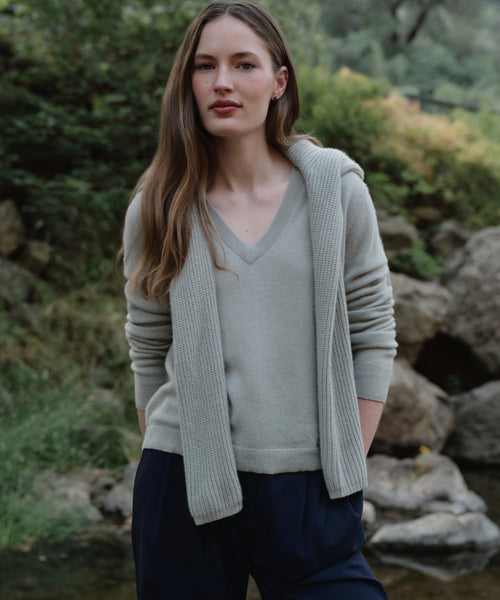 A woman with long brown hair stands outdoors by rocks and greenery, wearing the Flynn Cashmere Sweater in light gray, paired with a matching sweater draped over her shoulders and dark blue pants. She appears calm.