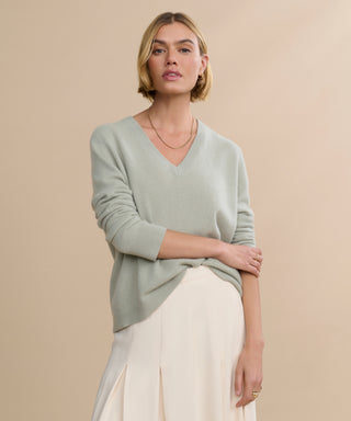 A woman with short blonde hair wears the Flynn Cashmere Sweater in pale green, styled with a cream skirt. She stands against a beige background, looking at the camera with a relaxed expression and one arm crossing her body.