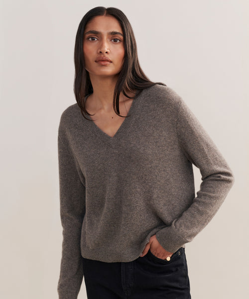 A woman with long, straight brown hair wears the Flynn Cashmere Sweater in a relaxed fit and black pants, standing against a plain light background and looking directly at the camera with a neutral expression.
