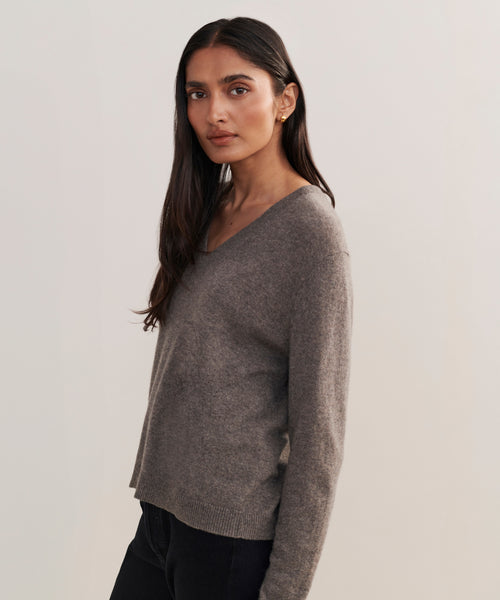 A woman with long dark hair stands against a plain light background, wearing the Flynn Cashmere Sweater—a relaxed fit knit V-neck—paired with black pants, looking slightly to the side with a neutral expression.