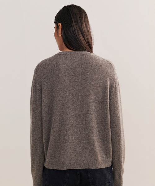 A person with long dark hair, seen from behind, wears the Flynn Cashmere Sweater in a relaxed fit grayish-brown knit with dark pants against a plain light background.