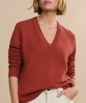 A person with short blonde hair wears the Flynn Cashmere Sweater in rust and light beige pants, standing against a plain beige background. The image is cropped at the shoulders and above the knees.
