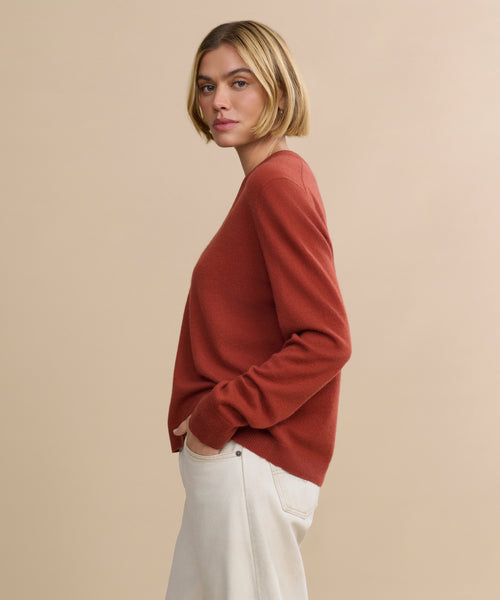 A woman with a blonde bob, wearing the Flynn Cashmere Sweater in rust and light beige pants, stands sideways with her hands in her pockets against a plain beige background.