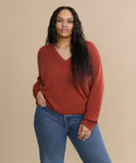 A woman with long wavy hair wears the Flynn Cashmere Sweater, a relaxed-fit knit V-neck in rust, paired with blue jeans. She stands against a plain beige background with one hand in her pocket.